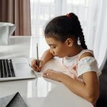 A girl focusing on homework, writing at a desk next to a laptop.