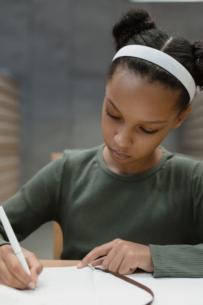 Young girl in a green sweater focusing on writing in a notebook indoors.
